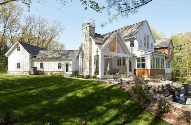Exterior view of a large, light-coloured multi-gabled house set in a mature wooded landscape. The home features white vertical siding, steep pitched gables, a prominent stone chimney, extensive windows, a covered entry porch, and a timber-toned glazed extension. A lawn, garden planting, and paved outdoor seating area surround the building.
