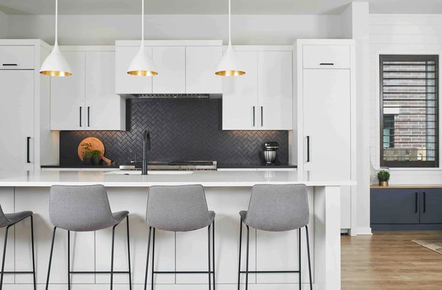 Bright contemporary kitchen with a white island and extensive white cabinetry, dark grey herringbone tile splashback, and three pendant lights with warm metallic interiors. Three grey upholstered bar stools line the island. A dark-framed window and additional dark lower cabinetry are visible to the right, with timber flooring throughout.