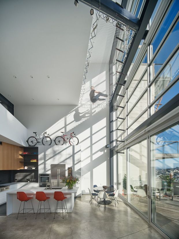 A bright, expansive contemporary kitchen and dining space with a dramatic double-height glazed wall. A suspended net lounge occupies the upper level, with a person sitting in it, while bicycles are mounted above the kitchen cabinetry. The kitchen features a white island with three red bar stools, dark cabinetry, timber wall panelling, stainless-steel appliances, and a polished concrete floor. A small round dining table sits beside the full-height glazing, which overlooks an urban landscape. Strong geometric shadows and abundant daylight create an airy, industrial-modern atmosphere.