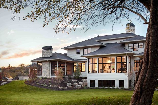 Exterior view of a large multi-level suburban home set in landscaped grounds. The house has a dark pitched roof with multiple gables, light stone masonry around the entry and chimney, pale horizontal siding, extensive black-framed windows, and a glazed projecting sunroom or living area. Mature trees and a gently sloping lawn surround the property.