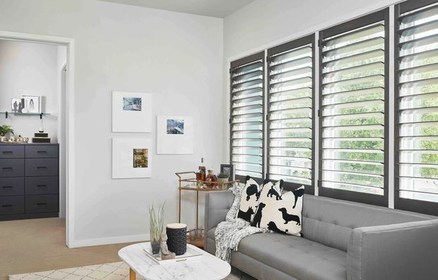 Bright contemporary living room with a grey upholstered sofa positioned beside a full-height bank of dark-framed louvred windows. The room has white walls, a neutral carpet, a small marble-look coffee table, a slim brass-toned drinks trolley, and restrained wall artwork. An adjoining room with dark built-in drawers is visible through the open doorway.
