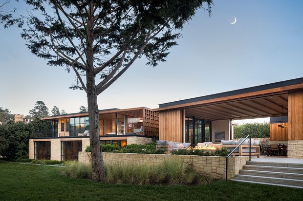 Exterior dusk view of a contemporary, low-slung residential house with extensive glazing, warm timber cladding and soffits, dark roof planes, and pale stone retaining walls. A covered outdoor entertaining area with seating and a linear fire feature opens onto landscaped grounds, with broad steps leading to the terrace. The composition also shows a mature tree, ornamental grasses, and a visible crescent moon.