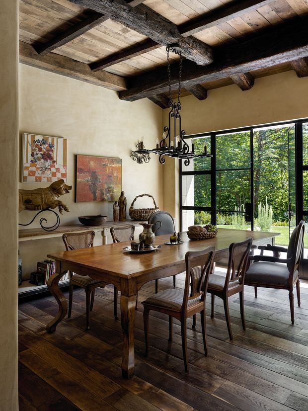 A warm, rustic dining room with a large solid timber dining table surrounded by woven-seat and upholstered chairs. The room features broad exposed dark timber ceiling beams, a timber plank ceiling and dark timber floorboards, textured cream plaster walls, an ornate wrought-iron chandelier, built-in shelving, and artwork. Large black-framed glazed doors or windows open the room visually to lush greenery outside.