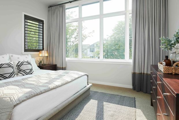 Bright, refined bedroom with a large white-framed multi-pane window, full-height grey curtains, a light upholstered bed with geometric black-and-white cushions, a dark timber bedside table and dresser, and a patterned grey area rug. The room has abundant natural light and a calm neutral palette.