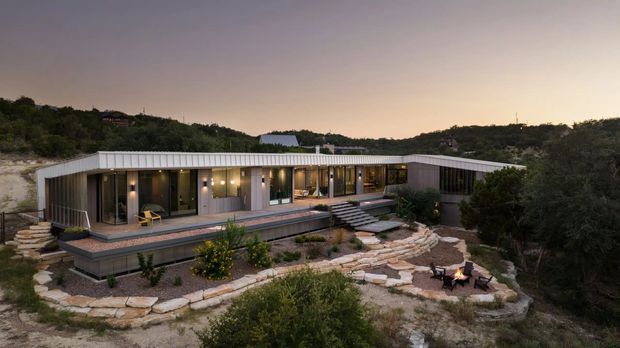 A low, single-storey contemporary residence set into a wooded, rocky landscape at dusk. The long horizontal building has a gently sloping metal roof, extensive floor-to-ceiling glazing, dark window frames, and vertical timber or wood-look cladding. A raised timber deck runs along the glazed facade, with steps leading down to layered stone paths and a small outdoor fire area in the foreground.