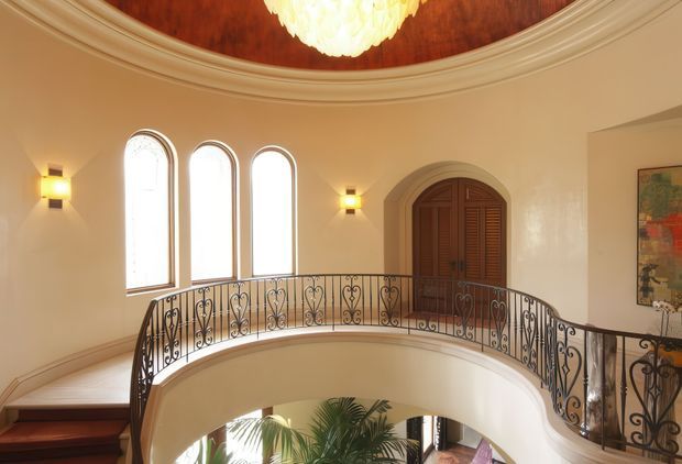 An elegant interior landing or upper-level gallery overlooking a lower floor, featuring a curved wrought-iron balustrade, three tall arched windows, an arched timber door with louvered panels, ornate ceiling mouldings, warm cream walls, wall sconces, and a central chandelier. A partial tropical plant and lower-level space are visible through the opening.