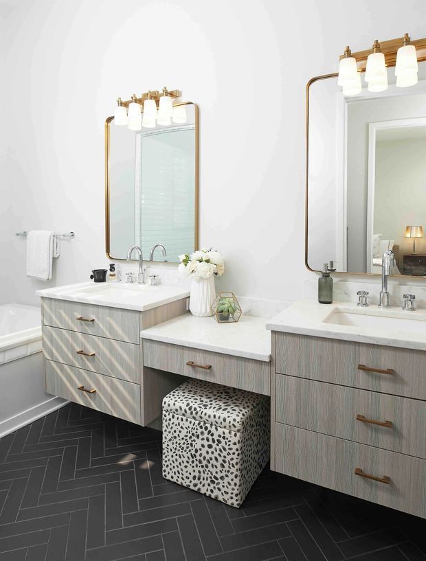 Bright, refined bathroom with a double vanity arrangement, two large rounded-rectangle mirrors, brass-toned vanity lighting and hardware, white benchtops, and pale vertically grained timber-look cabinetry. A freestanding-style bathtub is partially visible at left, while the dark charcoal herringbone floor creates contrast. A patterned upholstered stool and small decorative accessories add softness and character.