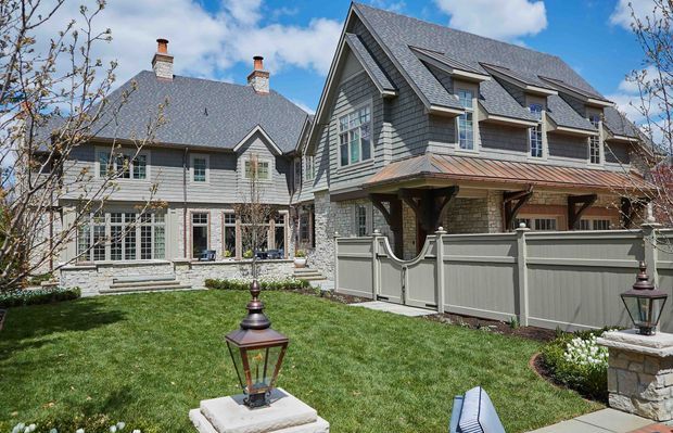 Exterior view of a large traditional multi-wing residence arranged around a landscaped courtyard. The home features steep gabled roofs with grey shingle roofing, grey shingle-style wall cladding, white-trimmed multi-pane windows, dormer windows, brick chimneys, and covered timber-supported entrances. A grey privacy fence, manicured lawn, stone edging, and lantern-style garden lights occupy the foreground.