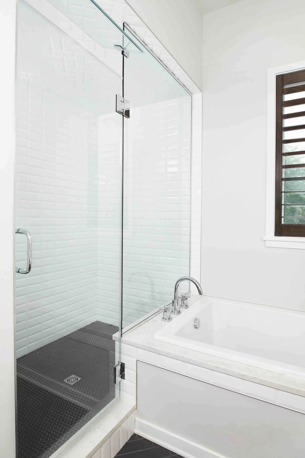 Bright contemporary bathroom featuring a large frameless glass walk-in shower with white textured wall tiles, dark small-format floor tiles, and a white built-in bathtub with chrome fixtures. A timber-framed louvred window provides natural light.