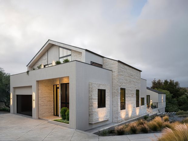 Exterior view of a contemporary multi-level residence with a strong gabled roof form, flat-roofed projecting volumes, pale textured masonry cladding, and vertically ribbed light-grey wall panels. Dark-framed windows and a recessed entry sit beside an integrated garage. The home is set on a landscaped, sloping site with ornamental grasses and paved areas, photographed in soft overcast evening light.