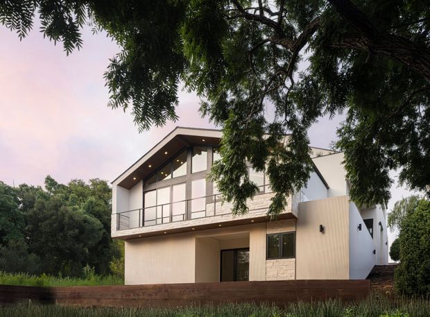 Exterior view of a contemporary two-storey house set among mature trees and landscaped grounds. The home has a strongly glazed upper-level facade with a projecting sloped roof, slim dark window and door frames, a balcony with metal balustrade, pale rendered walls, and a light stone-clad lower section. Warm recessed lighting is visible beneath the roof overhang and balcony.