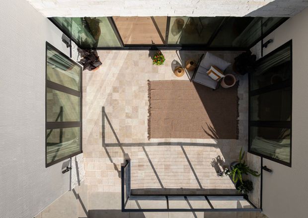 Top-down view of a bright enclosed courtyard or terrace surrounded by pale rendered walls and extensive black-framed glazing. The space has a light stone-paved floor, a large neutral outdoor rug, a grey lounge chair, potted plants, and strong geometric shadows from the surrounding glass and structure. The composition highlights an intimate indoor-outdoor living area with a clean contemporary feel.