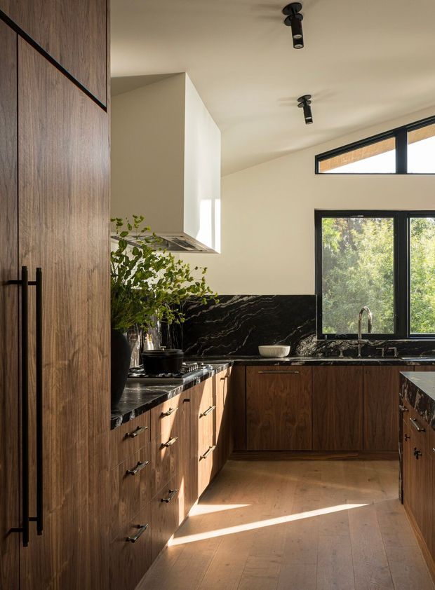 A warm, refined kitchen with dark timber cabinetry, black stone benchtops and a dramatic veined black stone splashback. A white sculptural rangehood contrasts with the cabinetry, while black-framed windows bring in abundant natural light and views of greenery. The space has timber flooring, streamlined black track lighting, and a compact galley/U-shaped layout.