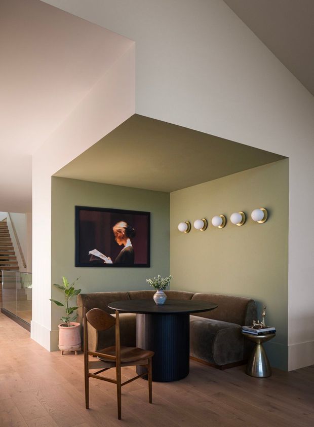 A refined open-plan living and dining nook beneath a dramatic angled ceiling. The space features a curved dark upholstered banquette, round black pedestal dining table, timber dining chair, wall-mounted television, sculptural wall lights, timber flooring, and muted sage-green and warm neutral walls. A staircase and glazed balustrade are visible at the left edge.