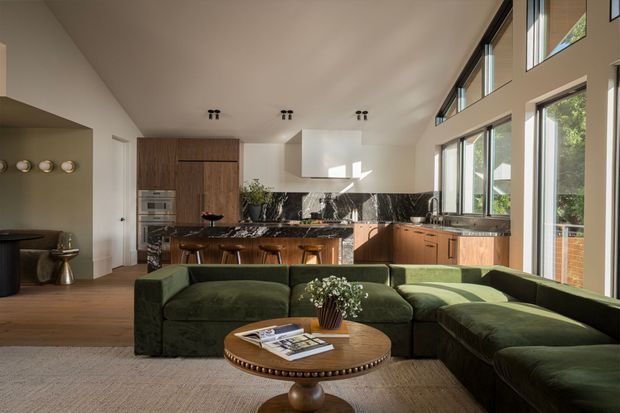 A bright open-plan living and kitchen area with a large deep-green sectional sofa in the foreground, a round timber coffee table, and a warm timber kitchen with a central island and bar seating. The kitchen features dark veined stone surfaces, extensive cabinetry, integrated appliances, and a white sculptural rangehood. Tall clerestory-style glazing and full-height windows bring in abundant natural light and overlook greenery. Warm timber flooring, neutral walls, and restrained contemporary detailing create a refined, inviting interior.