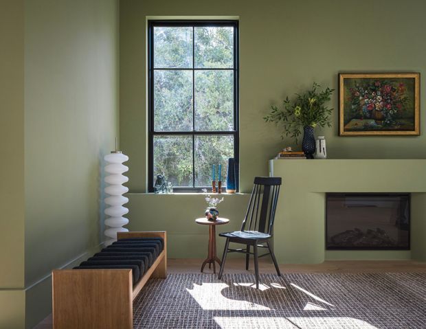 A compact, refined living room with muted sage-green walls, a tall black-framed multi-pane window, and a minimalist fireplace integrated into a projecting wall. The room contains a low timber lounge bench with dark cushions, a black spindle-back chair, a sculptural white floor lamp, a small side table, a vase arrangement, and framed floral artwork. Sunlight creates strong geometric patterns across the textured carpet.
