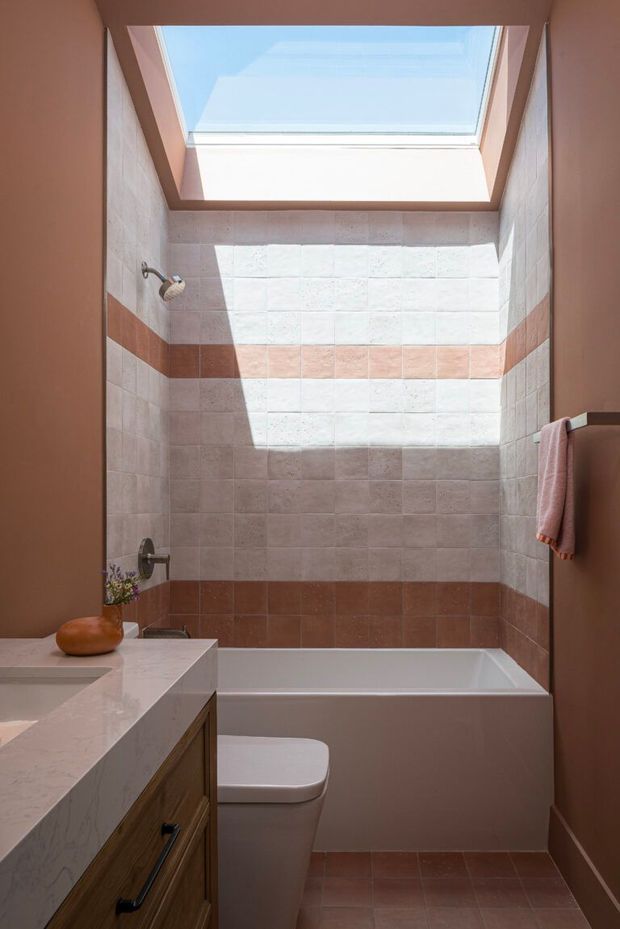 Compact bathroom with a bathtub and shower beneath a large sloped skylight. The walls are clad in pale rectangular tiles with horizontal terracotta accent bands and a darker terracotta tile base. A marble-look vanity with warm timber cabinetry occupies the foreground, accompanied by a white toilet. Natural sunlight creates a strong, warm, inviting atmosphere.