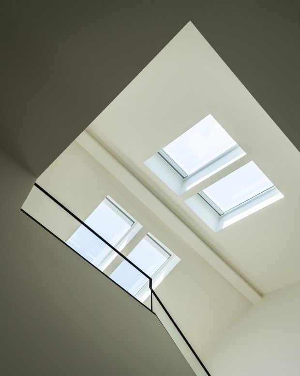 Upward-looking view of a bright, minimalist stairwell or landing with a high sloping ceiling, multiple rectangular skylights, and a slim black metal stair balustrade. The white architectural surfaces and abundant daylight create a clean, airy atmosphere.