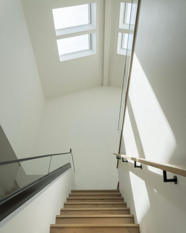 Bright modern stairway viewed from below, with pale timber treads, white plaster walls, a clear glass balustrade, and a timber handrail on black metal brackets. High-level windows and a skylight-like glazed opening flood the stairwell with natural light, creating a clean, airy vertical space.