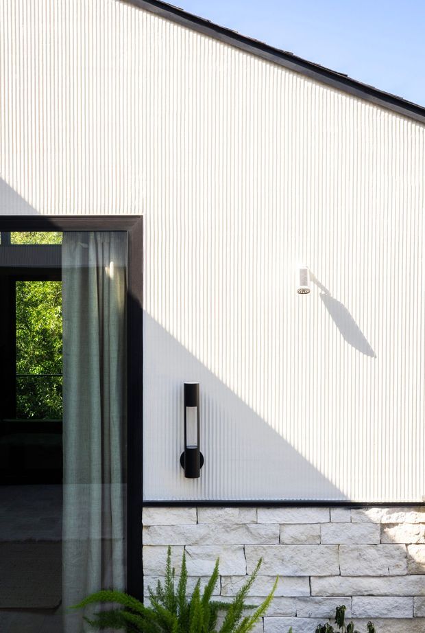 Close-up exterior view of a contemporary building facade. The wall is clad in vertically ribbed white material above a rough-cut pale stone base, with a dark metal-framed glazed opening and curtain at left, minimal black wall lights, and a low sloping roof edge. Strong sunlight creates crisp shadows across the facade.