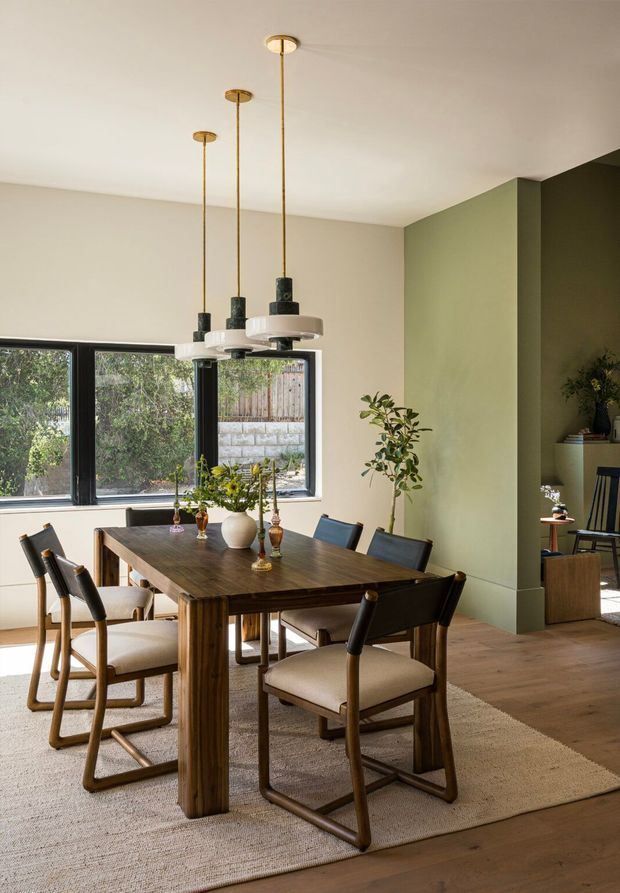 A warm, refined dining room centred on a substantial dark timber dining table with upholstered timber-framed chairs. Three suspended pendant lights hang at staggered heights above the table. The room has pale neutral walls, a muted sage-green architectural wall and dark green adjoining niche, timber flooring, a large natural-fibre rug, black-framed windows, and simple botanical styling. An adjoining seating or living area is partially visible through the open passage at right.