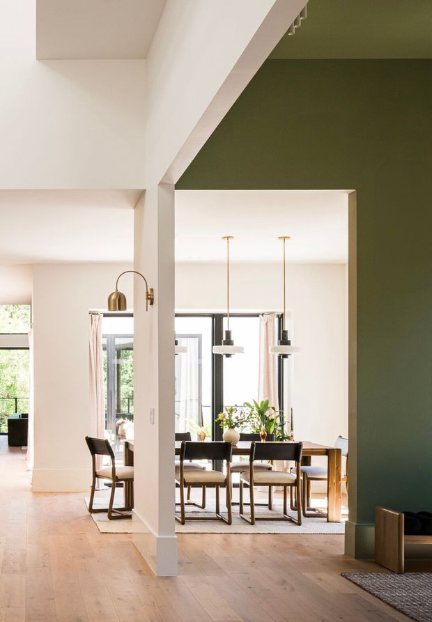 A bright, open-plan residential interior viewed from a circulation area, looking toward a dining room. The space features pale timber flooring, white walls and structural columns, a dark olive-green feature wall, a timber dining table with upholstered chairs, large glazed doors or windows, and two suspended pendant lights. The room has an airy, refined contemporary feel with strong architectural openings and generous natural light.