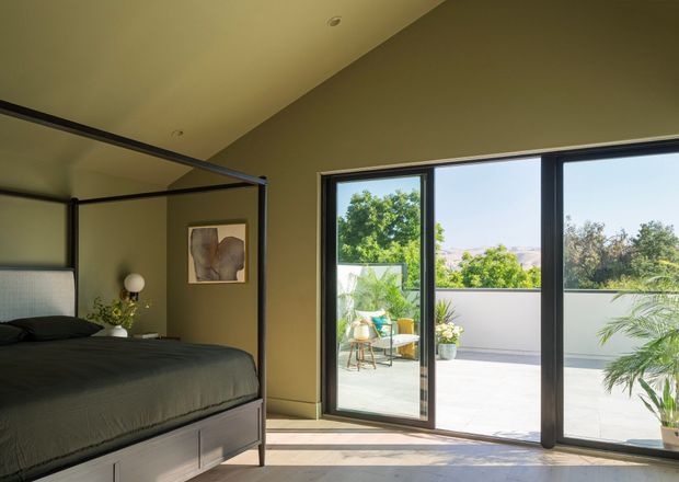A contemporary bedroom with a low platform bed and dark bedding, enclosed by a slim black-framed glass partition. Large black-framed sliding glass doors open onto a bright private terrace with outdoor seating and potted plants. The room has warm olive-grey walls, a pale ceiling, and abundant natural light.