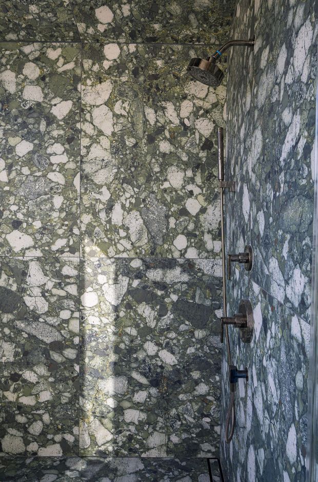 A close-up view of a luxurious walk-in shower clad almost entirely in dramatic green and pale veined natural stone slabs. A dark metal ceiling-mounted showerhead, handheld shower rail, controls, and fixtures are visible. The image is vertically oriented but presented rotated, with minimal detailing beyond the stonework and plumbing fixtures.
