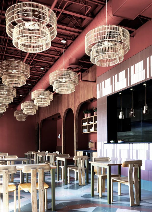Interior of a large hospitality venue, likely a restaurant or café, with a dramatic exposed industrial ceiling, prominent ductwork, suspended circular wire pendant chandeliers, deep burgundy walls, timber arches and shelving, a dark service counter, and rows of light timber tables and stools. The geometric tiled floor and tall clerestory-style windows add a bold, contemporary character.