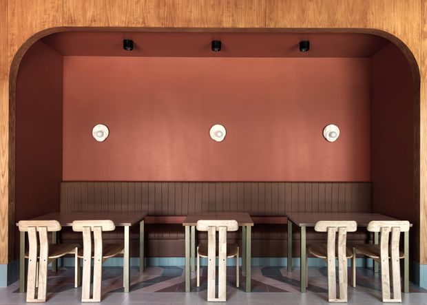 A compact, highly designed dining or café-style seating nook framed by warm timber panelling. The space features a deep terracotta-red wall and ceiling recess, a long dark wood vertical-ribbed banquette, small dark tables, and multiple pale timber stools. The floor has a geometric patterned finish in muted grey, blue, pink, and brown tones. Three round wall lights and small black ceiling spotlights provide illumination. The rounded timber arch and built-in seating create a refined contemporary hospitality-inspired interior.