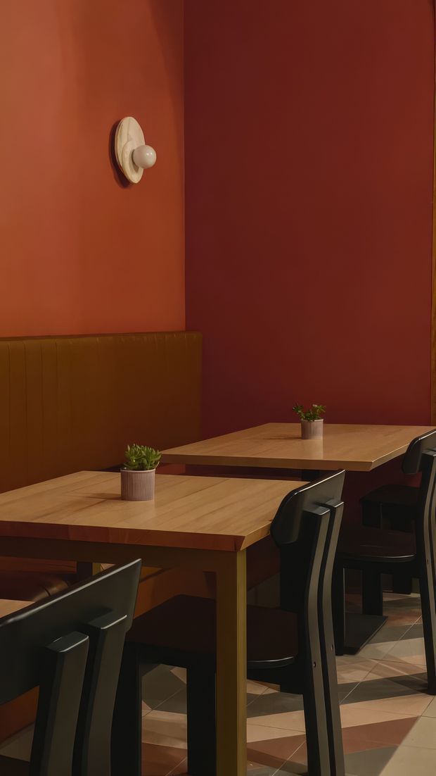 A moody, warmly lit dining area in a restaurant or café, with two timber tables, dark chairs, a built-in upholstered banquette, and small potted plants. The walls are painted in deep terracotta and rust tones, with a sculptural wall light. Geometric tiled flooring adds visual interest, creating an intimate contemporary setting.