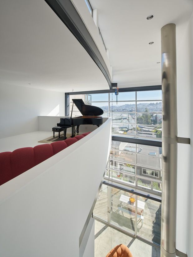 A bright, double-height interior overlooking a glazed atrium or performance space. A grand piano sits on an upper-level platform beside rows of curved red upholstered seating, separated by a white sculptural balustrade. Full-height glazing reveals an urban streetscape, while a large cylindrical structural column and exposed dark ceiling beam reinforce the contemporary public-building character.
