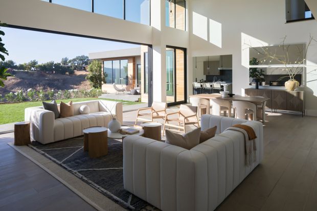 A bright, spacious open-plan contemporary interior with a living room, dining area, and kitchen connected by expansive floor-to-ceiling glazing. Two cream boucle-style sofas face a low coffee table and textured rug, while a round timber dining table and chairs sit near the kitchen. White walls, pale timber flooring, high ceilings, large glazed openings, and strong indoor-outdoor views create an airy, refined setting.