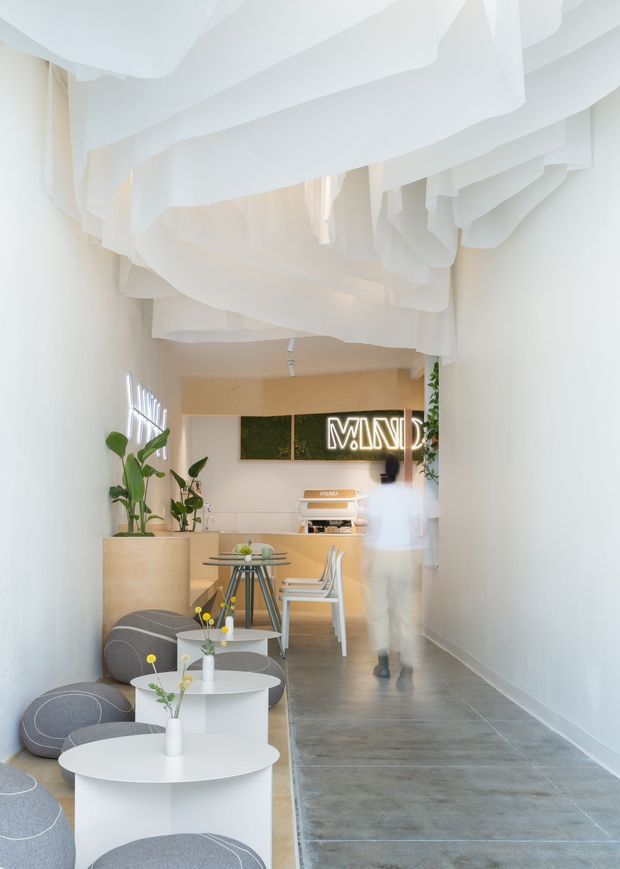 A bright, narrow contemporary commercial interior resembling a café, studio, or reception space. The room has a dramatic sculptural white fabric-like ceiling installation, smooth white walls, grey concrete-look tiled flooring, pale timber cabinetry, small round white tables, rounded grey upholstered seating, indoor plants, and a backlit sign. A blurred person is walking through the space.