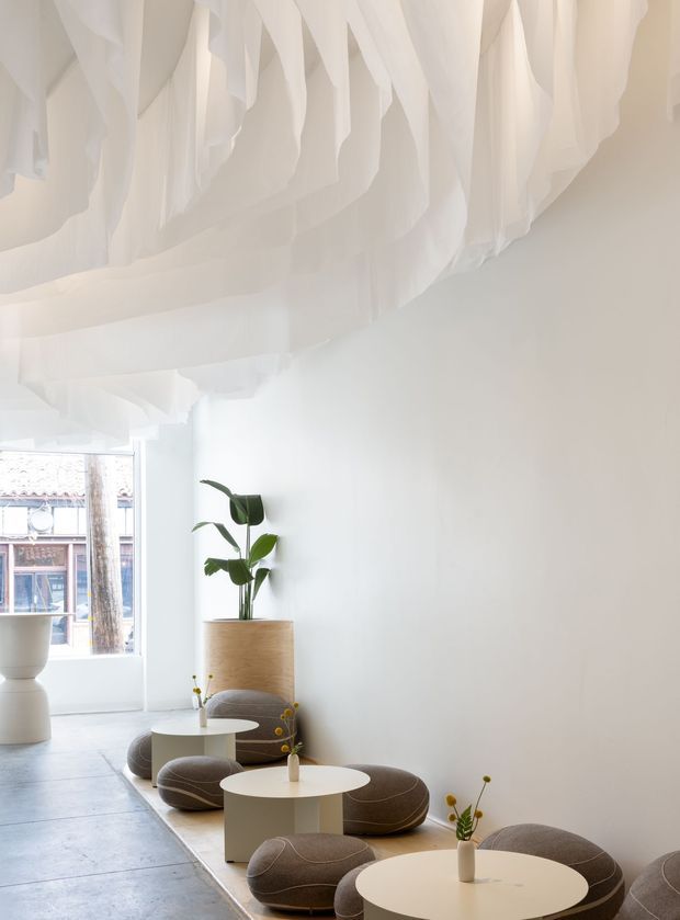 Bright minimalist interior with a sculptural installation of flowing white fabric suspended across the ceiling, white walls, pale tiled flooring, and a series of small round white tables paired with low rounded taupe upholstered stools. A large potted tropical plant adds greenery, while a street-facing window brings in natural light. The space resembles a refined café, lounge, or boutique commercial interior.