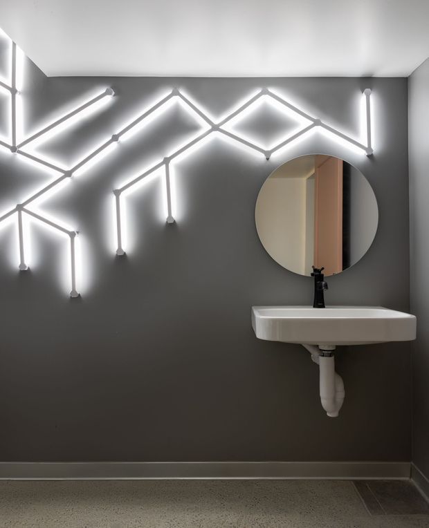A compact contemporary bathroom or powder room with dark charcoal-grey walls, a white wall-mounted rectangular basin and black tapware. A large round mirror hangs above the basin, while an expressive geometric arrangement of bright linear LED lights forms a branching pattern across the wall. The floor appears to be a speckled grey finish with a darker tiled perimeter or transition. The space has a dramatic, modern, high-contrast character.