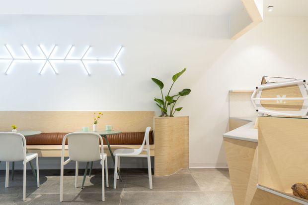 Bright contemporary commercial interior, likely a café or hospitality space, with a compact dining area, built-in timber banquette, pale dining chairs, a large potted plant, and a timber service counter or display joinery. The room has light walls, grey tiled flooring, a white sculptural wall light, and clean minimalist detailing.
