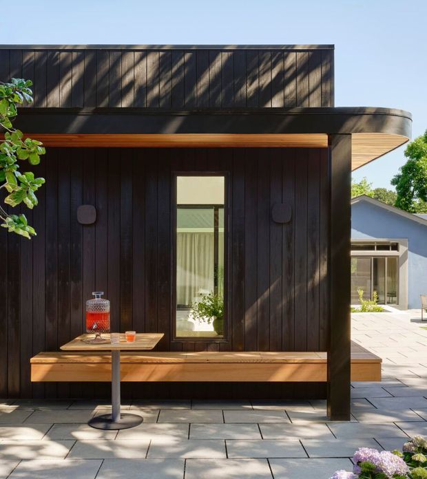 Exterior view of a contemporary dark timber-clad building with a flat roof and broad overhanging eaves. A built-in timber bench runs along the facade beside a narrow vertical window, with a small outdoor table and drink dispenser on a paved terrace. A lighter adjoining building and landscaped planting are visible in the background.
