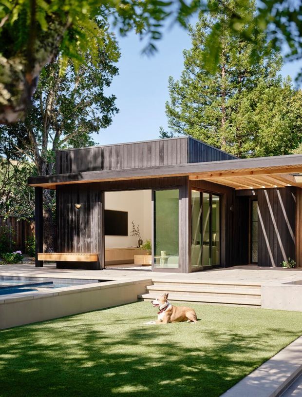 A modern single-storey house set within a leafy garden, featuring dark vertical timber cladding, a low-pitched roof, broad timber-framed glazing, and a covered outdoor deck or patio. The home opens onto a paved terrace and rectangular swimming pool, with a grass lawn in the foreground.
