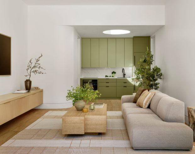 A bright, refined open-plan living room with a compact kitchen at the rear. The space features pale olive-green flat-panel cabinetry, a white marble-look splashback and benchtop, light timber flooring, a large neutral sectional sofa, woven coffee tables, a geometric area rug, a timber media console, and abundant indoor greenery. A circular ceiling skylight or light well adds natural illumination.