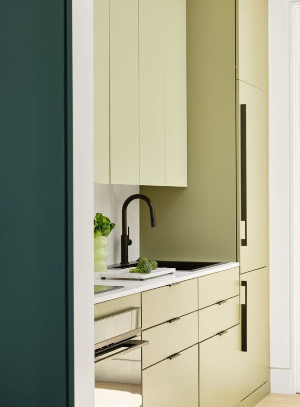 A compact contemporary kitchen viewed through a doorway, featuring pale sage-green full-height cabinetry, integrated appliances, slim dark handles, a white stone benchtop, dark splashback or wall surface, and a matte black gooseneck tap. A small open drawer reveals an integrated appliance, while a leafy plant and vegetables provide minor styling accents.