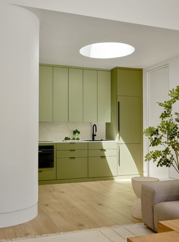 Bright open-plan kitchen viewed from an adjacent living area, featuring pale olive-green full-height cabinetry, integrated appliances, a light stone benchtop and splashback, black tapware, pale timber flooring, a curved white column, and a large oval ceiling skylight or rooflight. A neutral upholstered sofa and indoor tree are visible in the foreground.