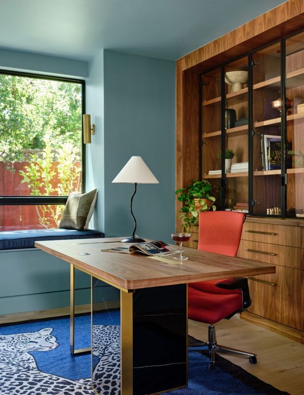 A stylish home office with a timber desk, built-in walnut-toned shelving and cabinetry, and black-framed glass cabinet doors. The room has blue-grey walls, a blue patterned rug, a red upholstered swivel chair, and a window seat beside a large garden-facing window. The design combines rich timber joinery with colourful mid-century-inspired furnishings and brass-toned accents.