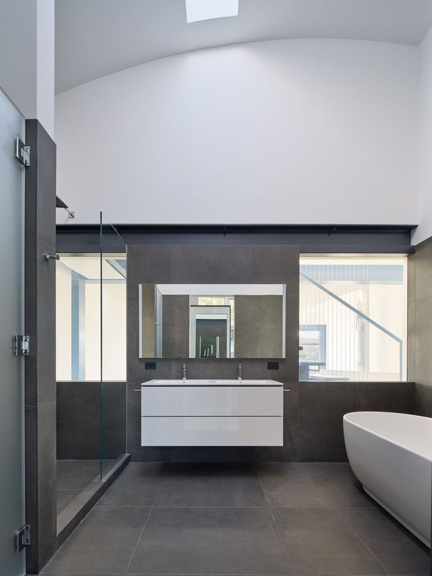 A contemporary bathroom with a freestanding white bathtub, wall-mounted double vanity, large rectangular mirror, and frameless glass shower enclosure. Dark grey tiled walls and floor contrast with the white fixtures. A high curved ceiling and skylight create an airy, bright interior, with glazing visible beyond the vanity.