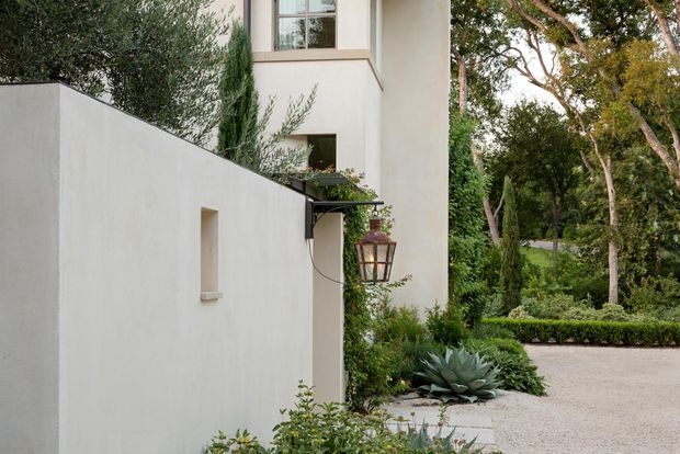 Exterior view of a refined contemporary residence, showing smooth pale rendered walls, a tall projecting building volume with large windows, and a dark metal wall-mounted lantern. A high rendered boundary wall frames a landscaped entry garden with sculptural succulents, clipped hedges, mature trees, and a pale gravel or paved driveway.