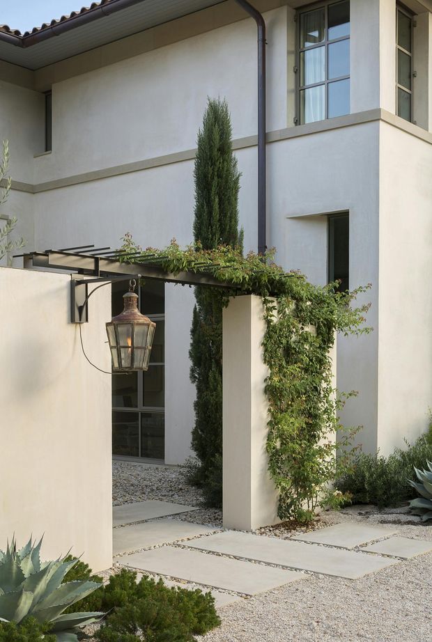 A refined courtyard entrance to a two-storey stucco residence. The pale rendered walls are paired with dark-framed divided-light windows, a tiled roof edge, and a slim dark downpipe. A simple pergola with climbing greenery frames the entry, which includes a hanging lantern, gravel landscaping, large rectangular stepping slabs, and sculptural drought-tolerant planting.