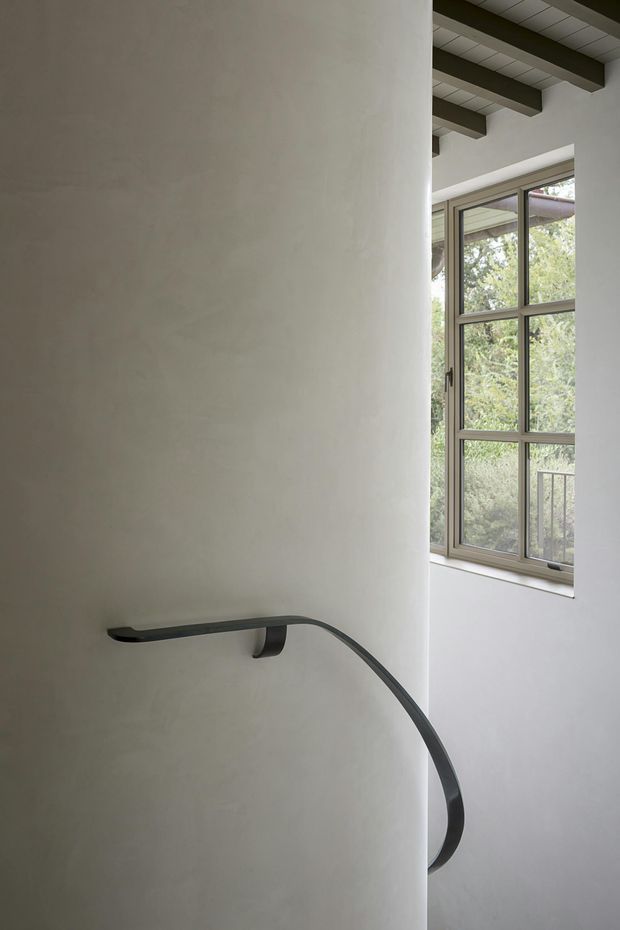 Interior architectural detail of a stairway with a sculptural dark metal handrail mounted against a smooth curved plaster wall. A tall multi-pane window brings in natural light and reveals greenery outside, while exposed dark ceiling beams and a gridded ceiling add structural character.