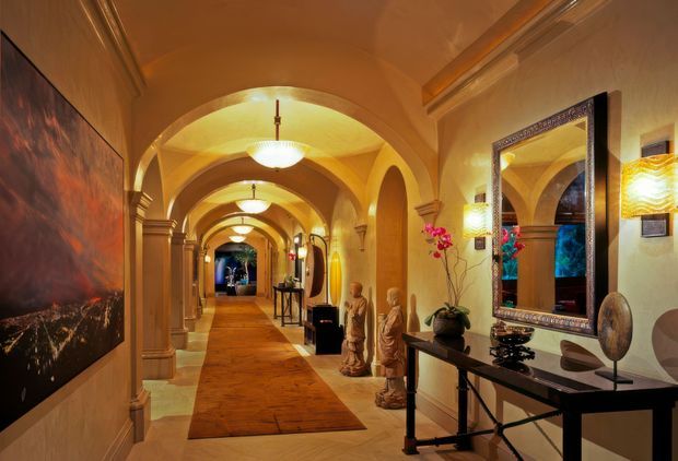 An ornate, warmly lit interior hallway with a long sequence of rounded arches, barrel-vaulted ceilings, decorative columns, and pendant lights. A patterned runner extends down the corridor, with console furniture, mirrors, sculptural statues, wall art, and decorative lighting along the sides. The space has a strongly Mediterranean-inspired, luxurious character.