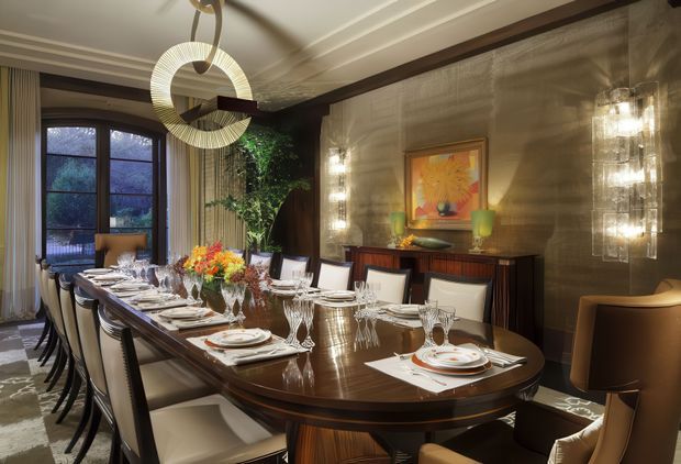 Elegant formal dining room centered on a long, highly polished dark wood dining table set for a large gathering. The room features upholstered dining chairs, layered table settings and glassware, a textured metallic-toned feature wall with artwork, sculptural pendant lighting, wall sconces, dark timber detailing, tall curtained windows, and indoor greenery. The overall atmosphere is luxurious, warm, and dramatic.