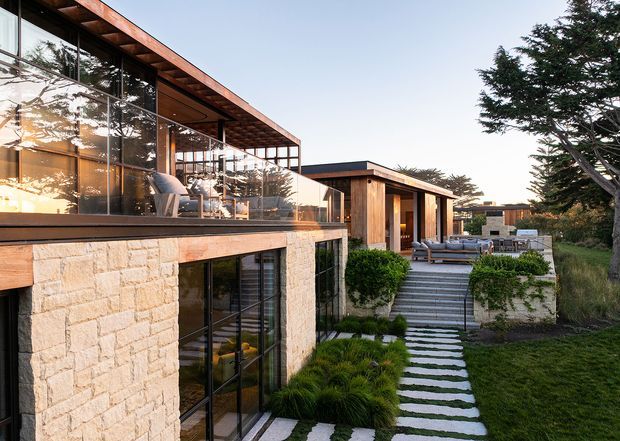 Exterior view of a modern multi-level residence integrated into a landscaped site. The house features extensive floor-to-ceiling glazing, warm timber soffits and cladding, pale natural stone walls, broad horizontal rooflines, and outdoor terraces connected by stepped stone pathways. A furnished outdoor lounge is visible on the upper terrace, with established coastal-looking trees and grasses surrounding the building.